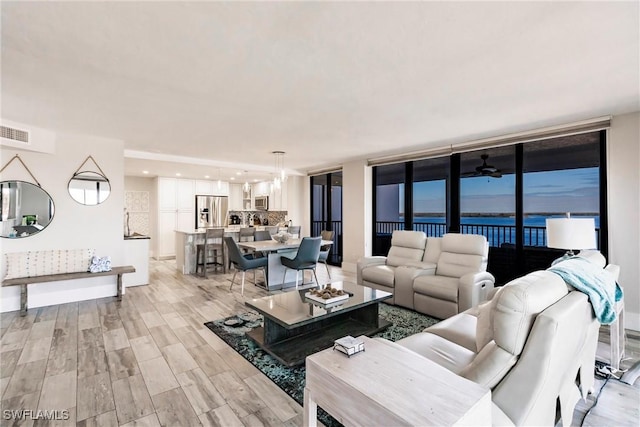 living room featuring floor to ceiling windows, ceiling fan, a water view, and light wood-type flooring