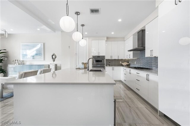 kitchen featuring sink, wall chimney exhaust hood, hanging light fixtures, an island with sink, and white cabinets