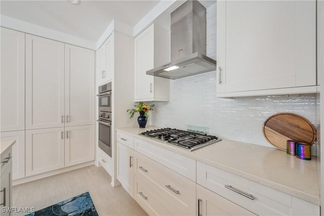 kitchen with wall chimney range hood, tasteful backsplash, light stone counters, white cabinets, and appliances with stainless steel finishes