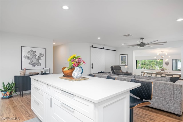 kitchen with ceiling fan with notable chandelier, a kitchen island, a barn door, light hardwood / wood-style flooring, and white cabinetry