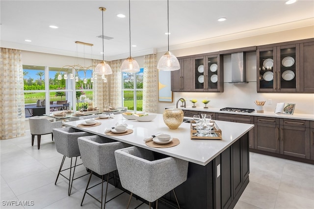 kitchen featuring stainless steel gas stovetop, a kitchen island with sink, a kitchen breakfast bar, wall chimney range hood, and decorative light fixtures