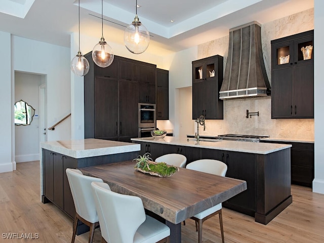 kitchen with pendant lighting, a center island with sink, wall chimney exhaust hood, light wood-type flooring, and a kitchen bar