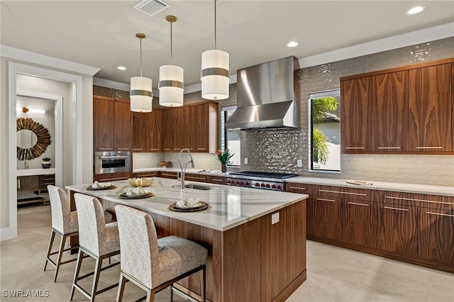 kitchen with appliances with stainless steel finishes, sink, wall chimney range hood, a center island with sink, and hanging light fixtures