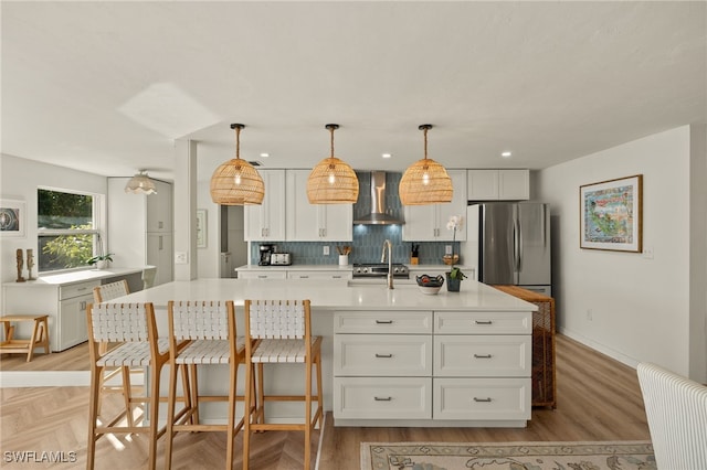 kitchen with stainless steel refrigerator, white cabinetry, wall chimney range hood, an island with sink, and decorative light fixtures