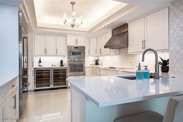 kitchen with stainless steel double oven, a tray ceiling, wine cooler, wall chimney exhaust hood, and backsplash