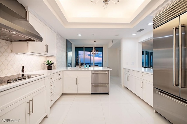 kitchen featuring a tray ceiling, black electric stovetop, stainless steel built in fridge, wall chimney range hood, and a chandelier