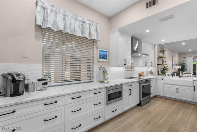 kitchen with light stone countertops, wall chimney range hood, appliances with stainless steel finishes, white cabinets, and light wood-type flooring