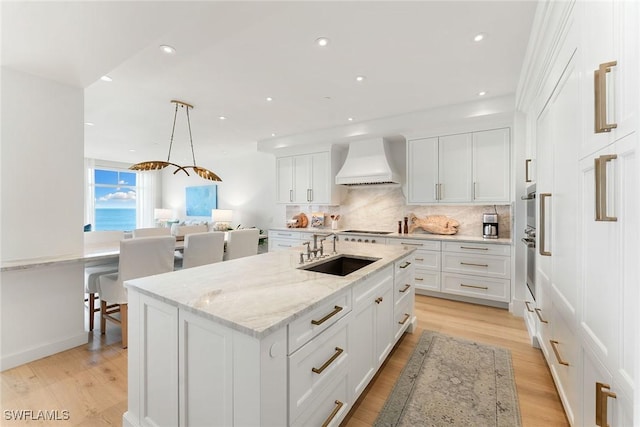 kitchen with an island with sink, sink, white cabinets, custom exhaust hood, and hanging light fixtures