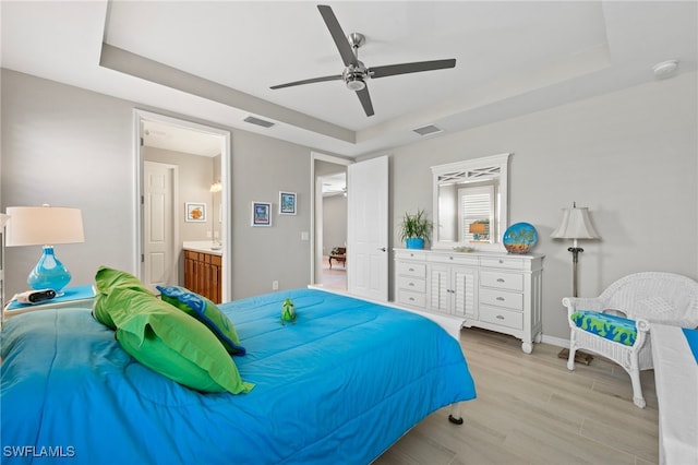 bedroom with ceiling fan, light hardwood / wood-style floors, ensuite bathroom, and a tray ceiling