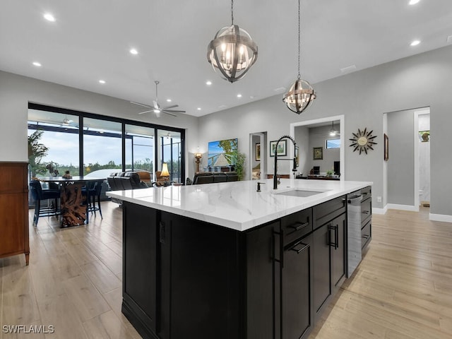 kitchen with sink, hanging light fixtures, light stone counters, a center island with sink, and ceiling fan with notable chandelier