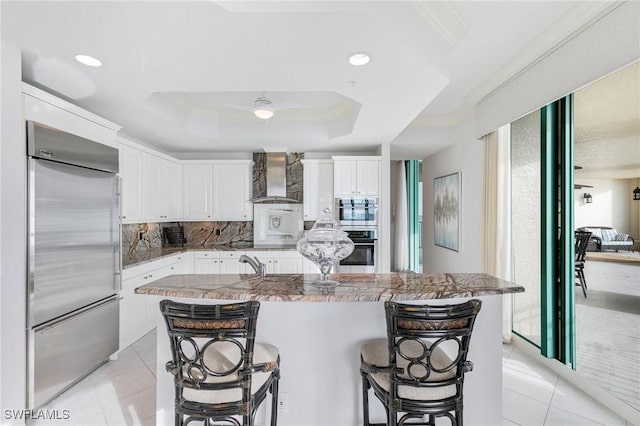 kitchen with a breakfast bar area, light tile patterned floors, a tray ceiling, stainless steel appliances, and wall chimney range hood