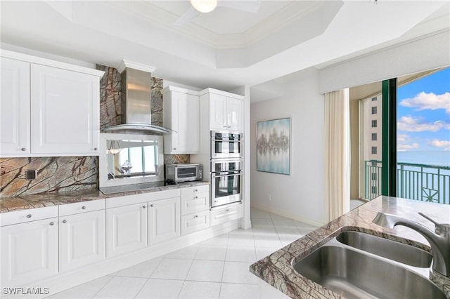 kitchen featuring a tray ceiling, black electric stovetop, a sink, double oven, and wall chimney range hood