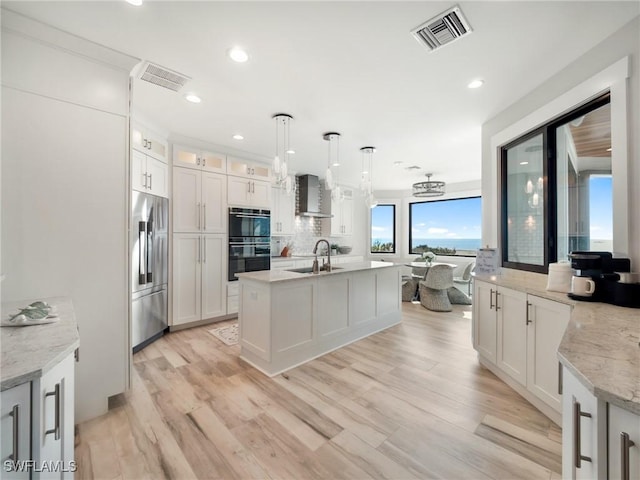 kitchen featuring wall chimney exhaust hood, light stone counters, stainless steel built in refrigerator, decorative light fixtures, and white cabinets