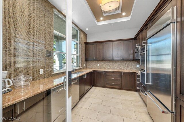 kitchen featuring sink, a tray ceiling, stainless steel appliances, light stone countertops, and decorative backsplash