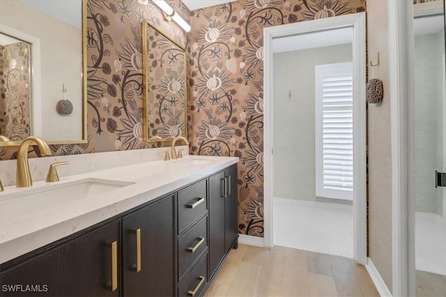 bathroom featuring wallpapered walls, a sink, double vanity, and wood finished floors