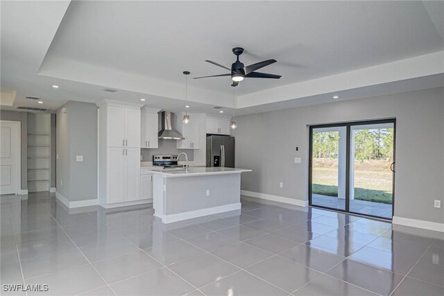 kitchen featuring appliances with stainless steel finishes, an island with sink, white cabinets, a tray ceiling, and wall chimney range hood