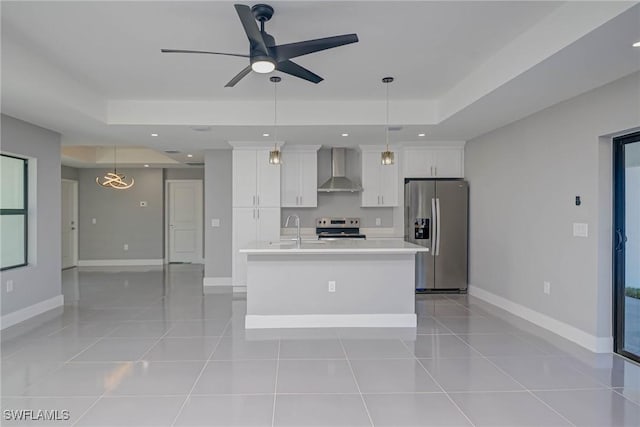 kitchen with wall chimney range hood, white cabinetry, a kitchen island with sink, stainless steel appliances, and a tray ceiling