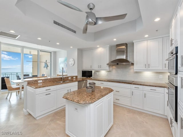 kitchen with a raised ceiling, a kitchen island, and wall chimney range hood