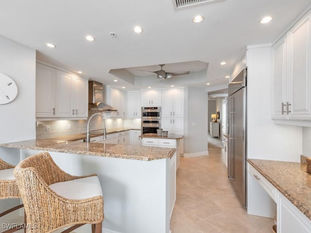 kitchen with kitchen peninsula, light stone countertops, wall chimney range hood, white cabinets, and a breakfast bar area