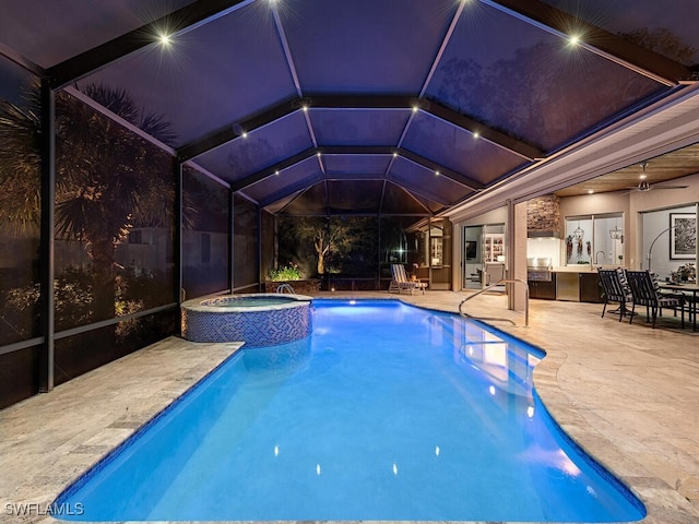 pool at night with glass enclosure, ceiling fan, an outdoor kitchen, an in ground hot tub, and a patio