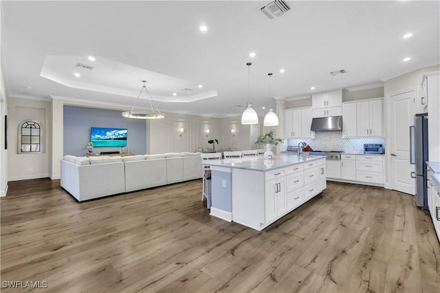 kitchen featuring decorative light fixtures, white cabinetry, decorative backsplash, a kitchen island with sink, and a tray ceiling