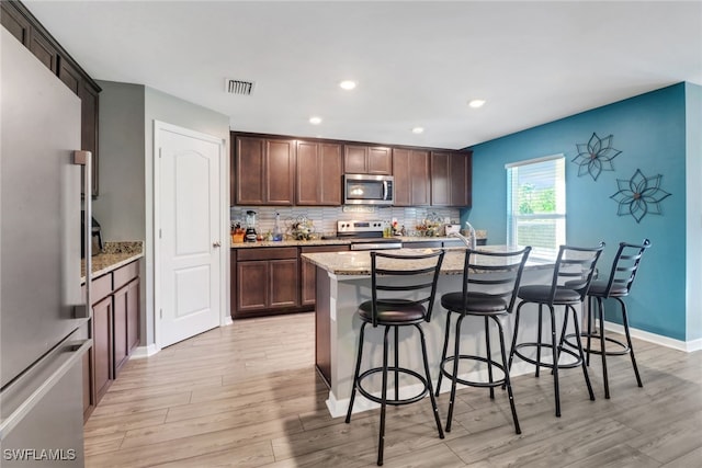 kitchen with a kitchen island with sink, light hardwood / wood-style flooring, dark brown cabinets, light stone counters, and stainless steel appliances