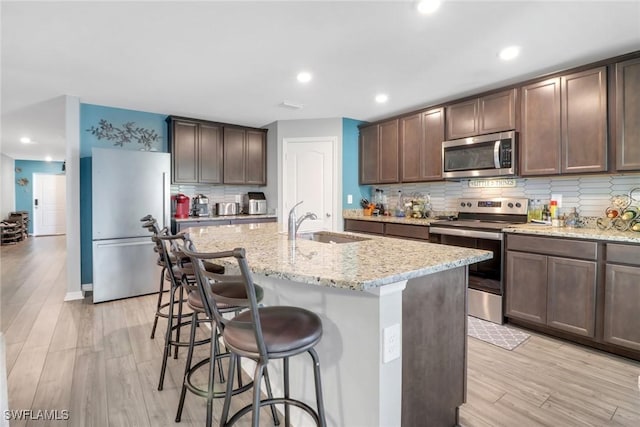 kitchen with sink, an island with sink, appliances with stainless steel finishes, dark brown cabinets, and light stone counters