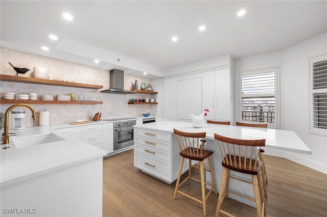 kitchen with stainless steel oven, wall chimney exhaust hood, sink, light hardwood / wood-style flooring, and white cabinets