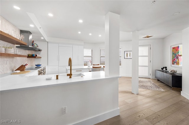 kitchen with sink, wall chimney range hood, kitchen peninsula, white cabinets, and light wood-type flooring