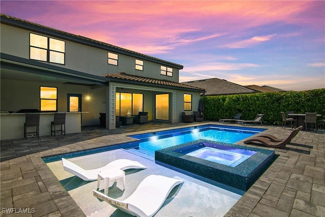 pool at dusk with an in ground hot tub, a patio area, and exterior bar