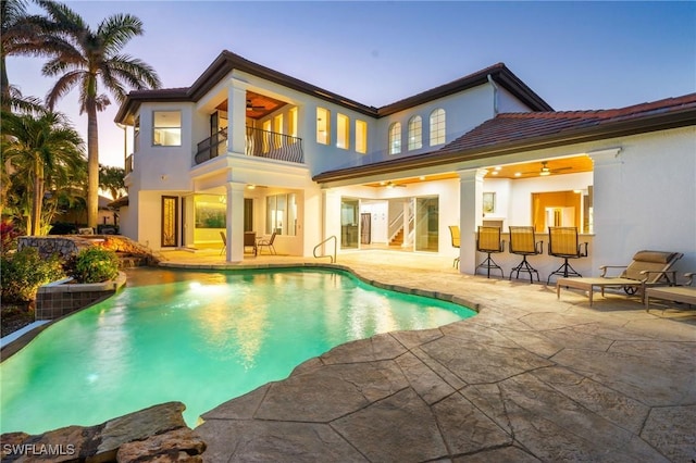 back house at dusk featuring ceiling fan, a balcony, an outdoor bar, and a patio