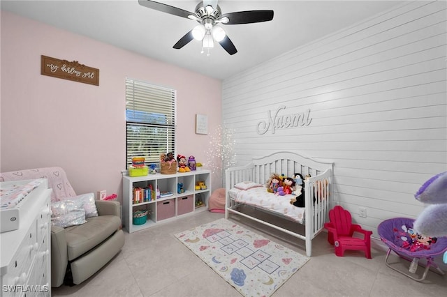 bedroom with ceiling fan, wood walls, light tile patterned floors, and a crib