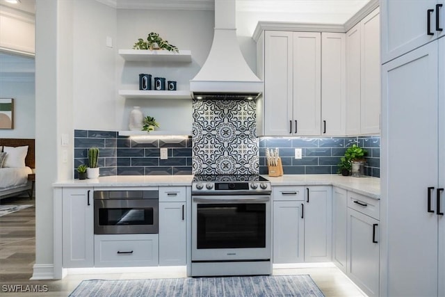 kitchen featuring stainless steel electric stove, premium range hood, tasteful backsplash, light stone countertops, and light hardwood / wood-style floors