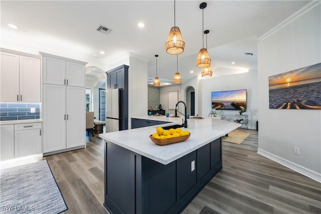 kitchen with fridge, tasteful backsplash, white cabinetry, decorative light fixtures, and an island with sink