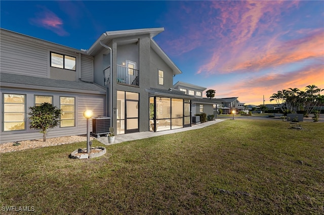 back house at dusk featuring a sunroom, central AC unit, a balcony, and a yard