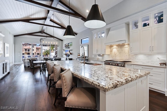 kitchen with white cabinets, decorative light fixtures, premium range hood, and a kitchen island with sink