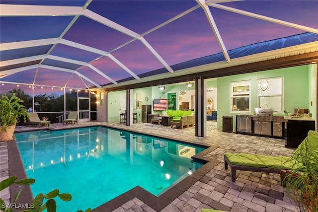 pool at dusk featuring outdoor lounge area, a patio, an outdoor kitchen, and glass enclosure