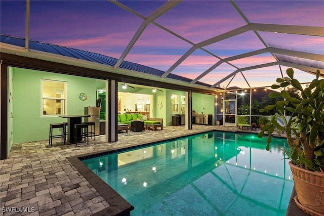 pool at dusk with an outdoor living space, glass enclosure, exterior bar, and a patio