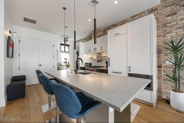 kitchen with brick wall, sink, pendant lighting, white cabinetry, and an island with sink