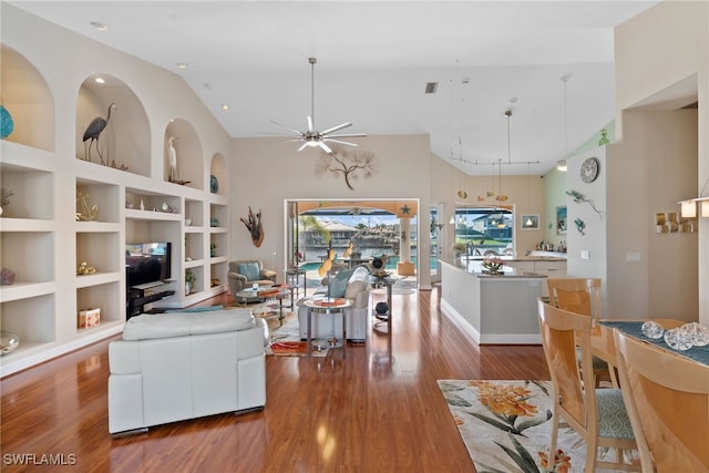 living room with built in shelves, ceiling fan, a towering ceiling, and wood-type flooring