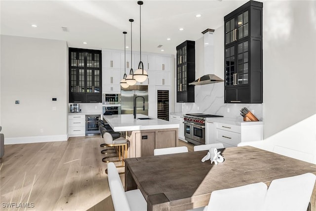 kitchen with wall chimney exhaust hood, premium appliances, a kitchen island with sink, pendant lighting, and white cabinets