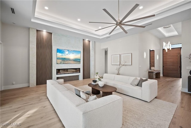 living room featuring a raised ceiling, a large fireplace, ceiling fan with notable chandelier, and light wood-type flooring