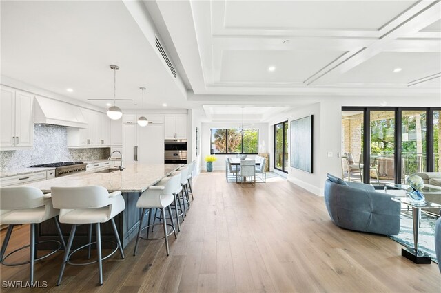 kitchen with light stone countertops, custom range hood, a large island with sink, white cabinetry, and hanging light fixtures