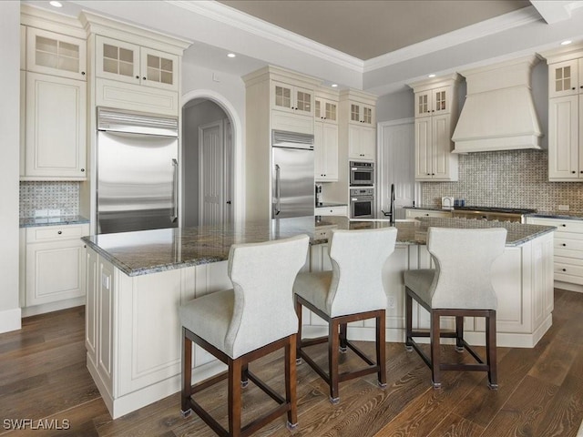 kitchen featuring dark wood-type flooring, premium range hood, a large island with sink, dark stone counters, and appliances with stainless steel finishes