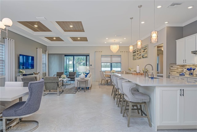 kitchen featuring decorative light fixtures, sink, coffered ceiling, an island with sink, and white cabinets