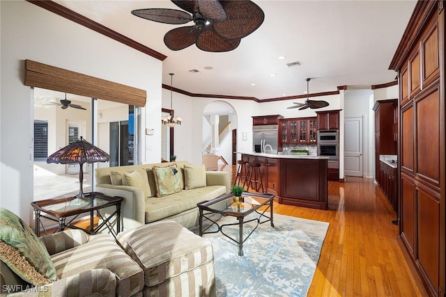 living area featuring arched walkways, crown molding, recessed lighting, light wood-style floors, and ceiling fan with notable chandelier