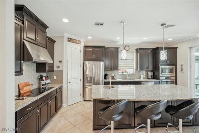 kitchen with decorative light fixtures, stainless steel appliances, an island with sink, ornamental molding, and sink