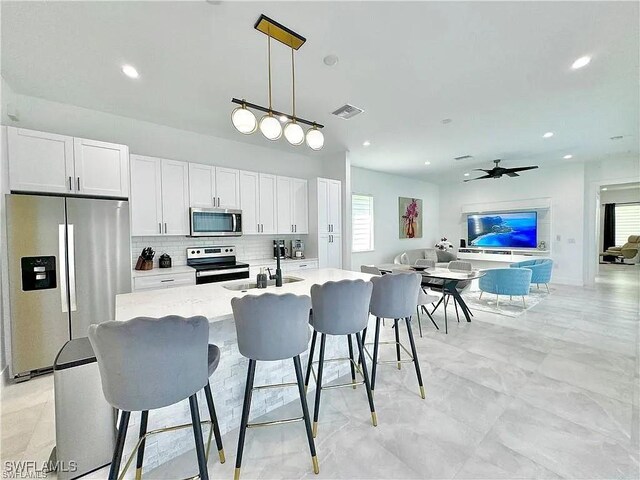 kitchen with appliances with stainless steel finishes, ceiling fan, pendant lighting, a center island with sink, and white cabinets