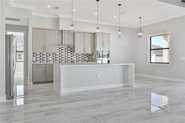 kitchen with gray cabinets, hanging light fixtures, a center island with sink, and wall chimney range hood