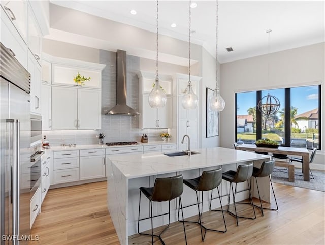 kitchen with wall chimney exhaust hood, an island with sink, decorative backsplash, white cabinets, and sink